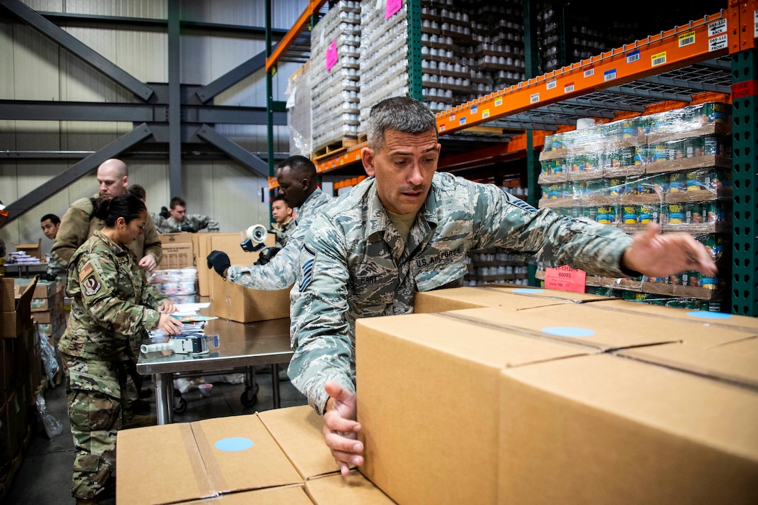 California Air National Guardsmen handle cardboard boxes and food products in a large, warehouse-type room.