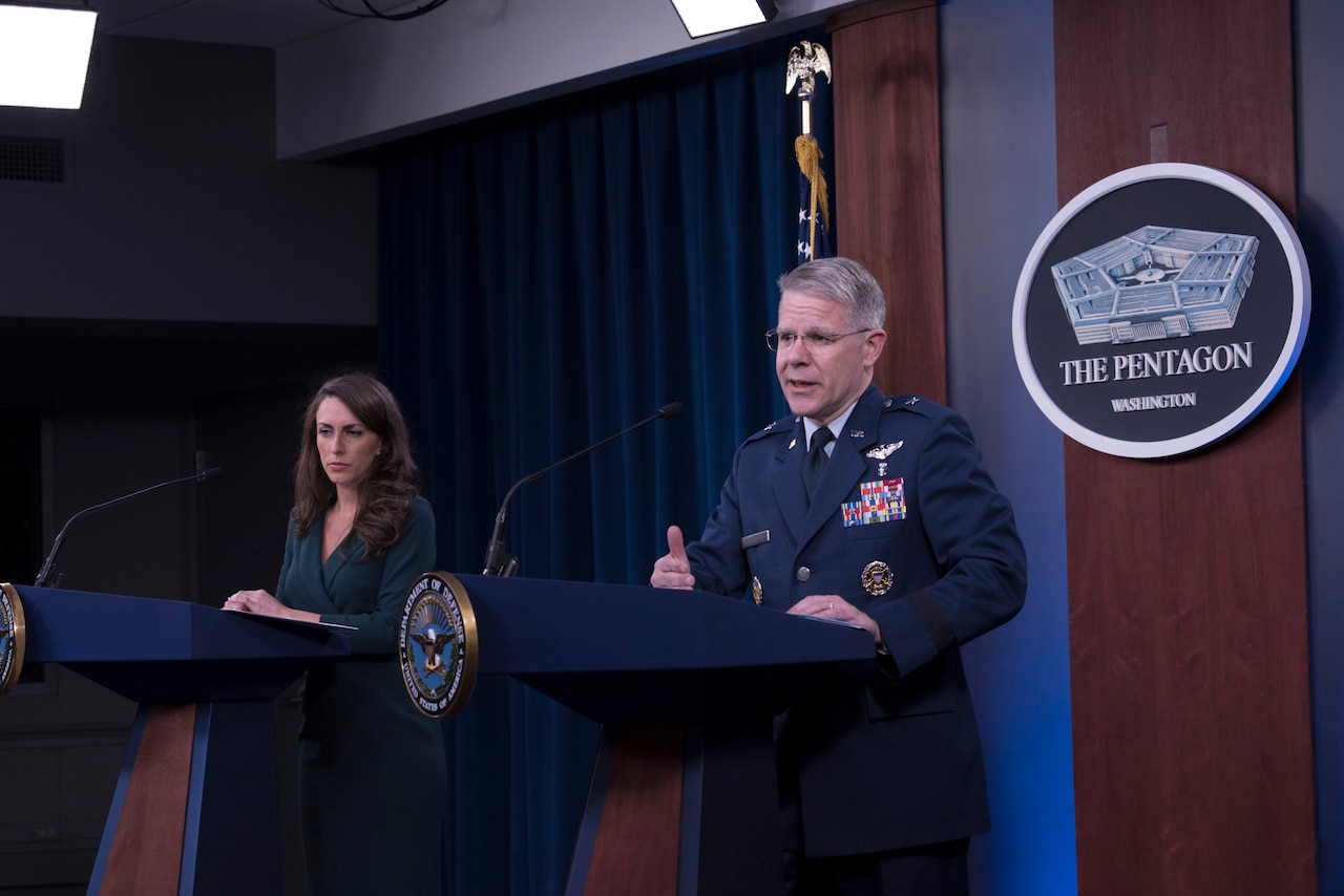 A woman in civilian clothes and a man in uniform stand behind lecterns.