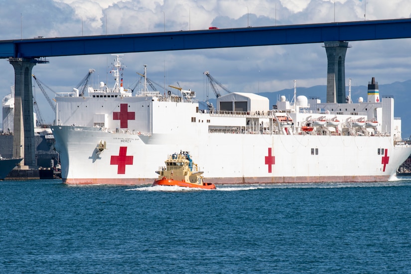 A Navy ship with a red cross on its side is escorted by a small tug boat near a bridge.