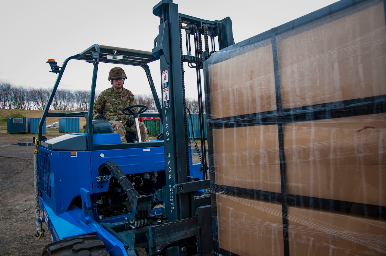 A military member operates a forklift to move a palette of boxes.