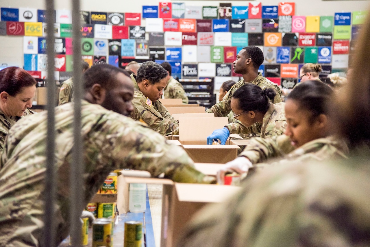 Service members wearing gloves put food into boxes.