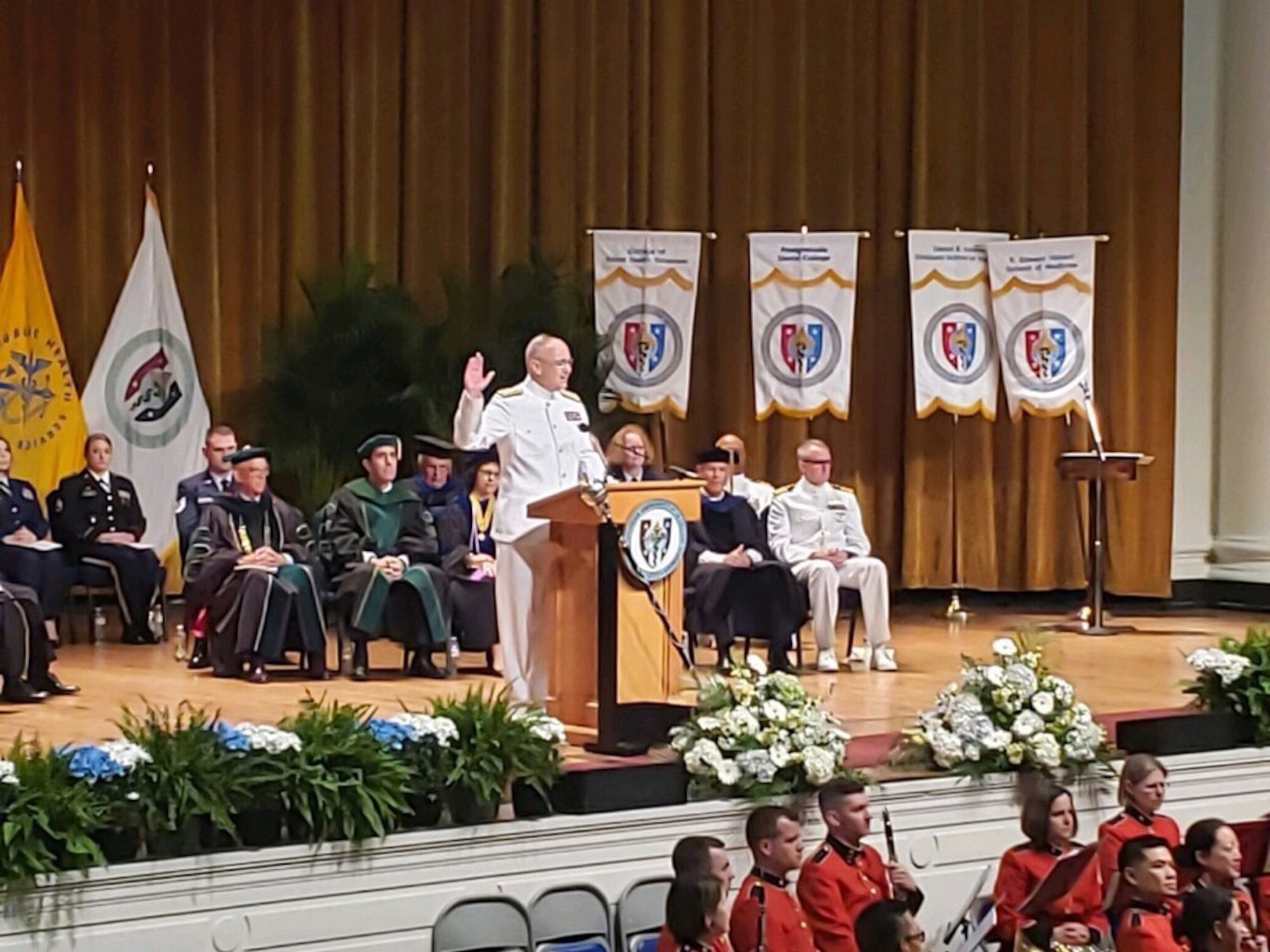 Navy admiral holds up his right hand to administer an oath while standing behind a lectern on an auditorium stage.