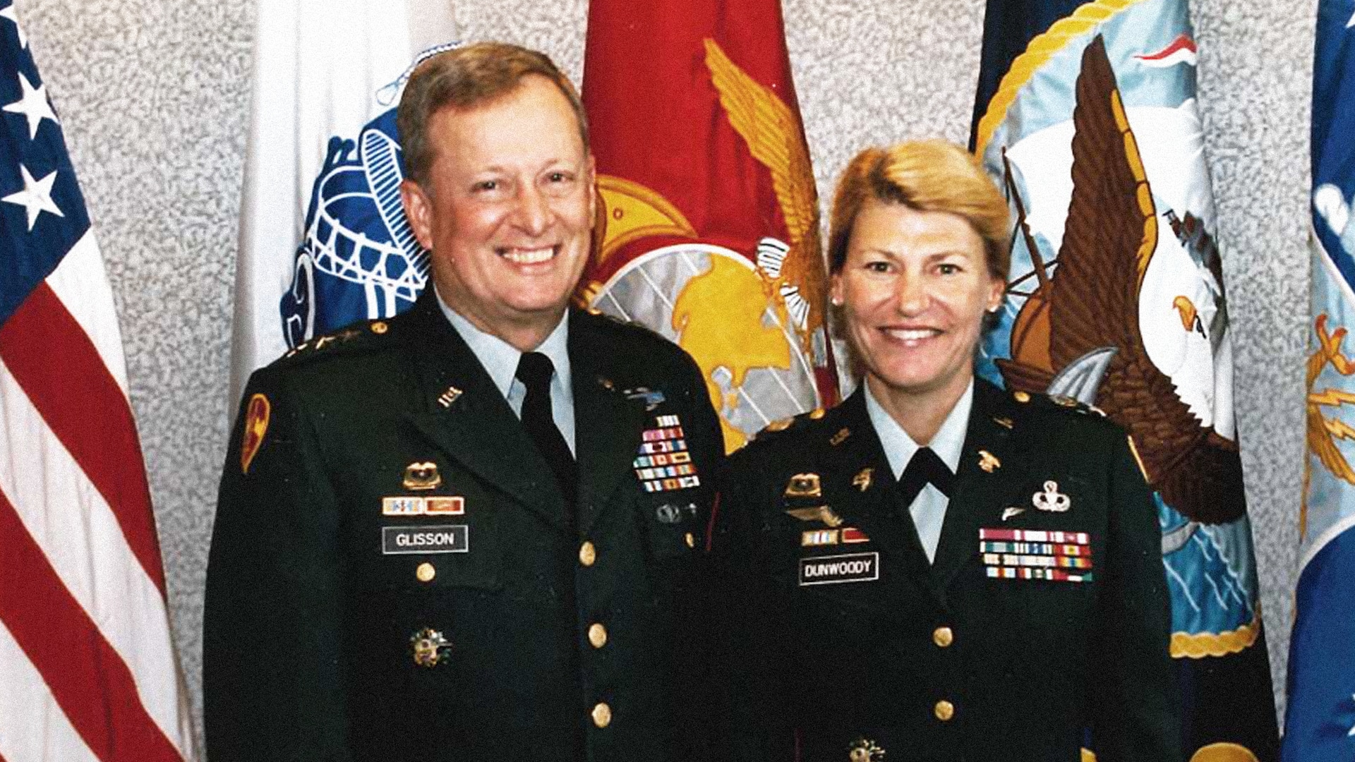 Male and female officer in Army dress green uniform stand in front of flags.