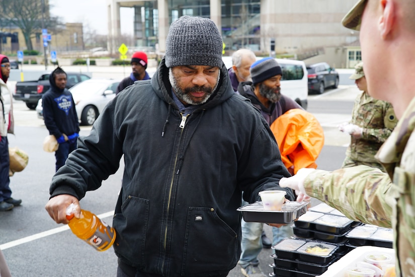 Soldier hands a packaged hot meal to a civilian.