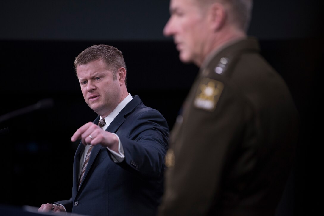 Man at lectern gestures and man in military uniform looks on.
