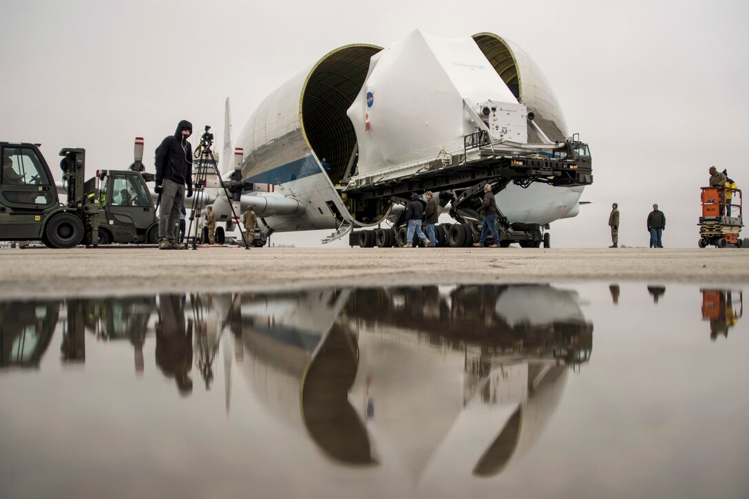 Airmen and other personnel mill about an open aircraft and a space capsule on a flightline.