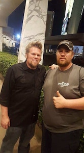 One male with blond hair and beard in black shirt and blue jeans stands next to man with beard in grey shirt, baseball cap, and black pants making a thumbs up sign.