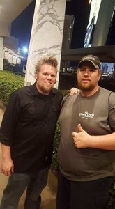 One male with blond hair and beard in black shirt and blue jeans stands next to man with beard in grey shirt, baseball cap, and black pants making a thumbs up sign.