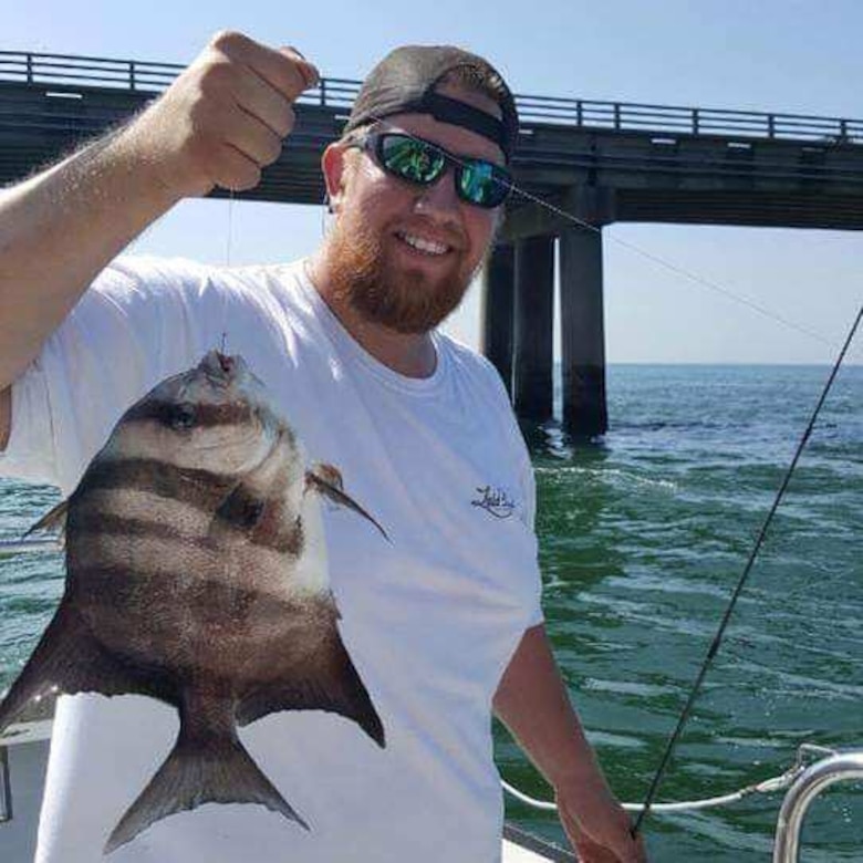 White male with beard in white shirt, sunglasses, baseball cap backwards holds up a a grey and white striped fish.