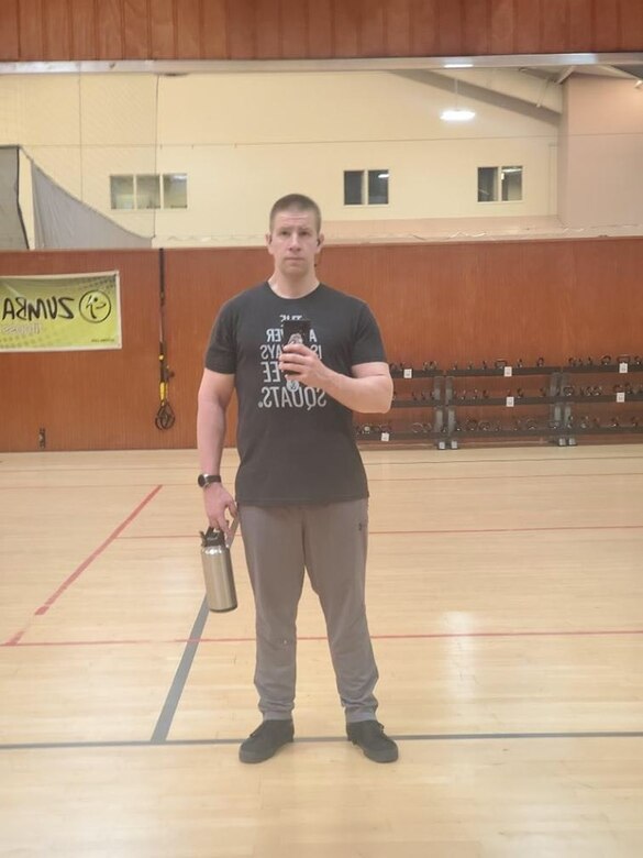 Man in black and white t-shirt and gray pants and black shoes holds phone in front of his chest in left hand and a stainless steel water bottle in right hand.
