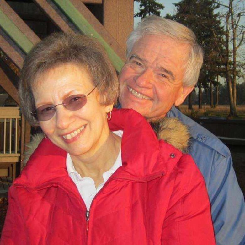 Woman in red coat, white shirt, glasses, and small hoop earrings stands in front of a man with white hair in blue coat outside of a house.