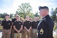 Group of young men and women in black shirts and kaki pants stand outside as man in Army blue dress uniform and beret stands to the front of their formation.