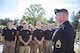 Group of young men and women in black shirts and kaki pants stand outside as man in Army blue dress uniform and beret stands to the front of their formation.