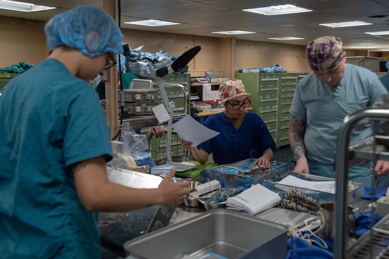 Sailors wearing surgical scrubs prepare equipment for sterilization.