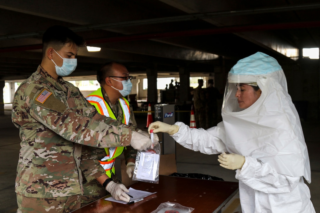 A soldier in a white protective outfit places a sample into a bag that another soldier is holding.