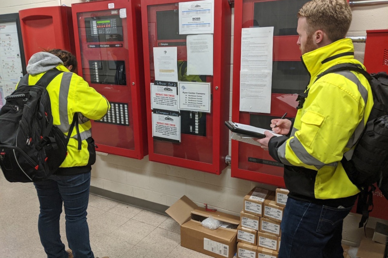 A woman and a man, both wearing yellow coats and black backpacks, make notes on clipboards.