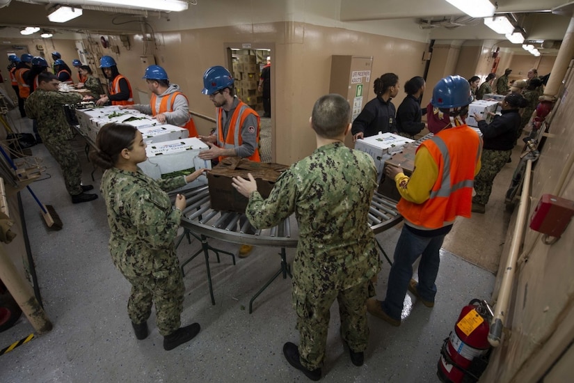 Sailors move boxes of supplies along a roller track.