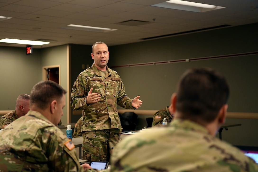 Standing airman gestures while speaking to airmen seated at tables.