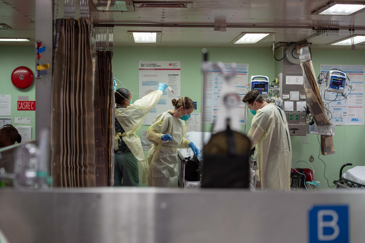 Three sailors wearing masks and gloves tend to a patient in a hospital bed.