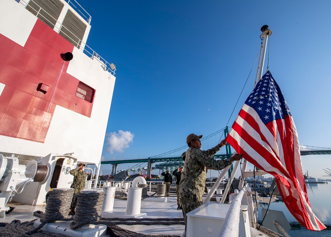 Sailor raises flag on ship marked with a large red cross as another sailor salutes in the background.