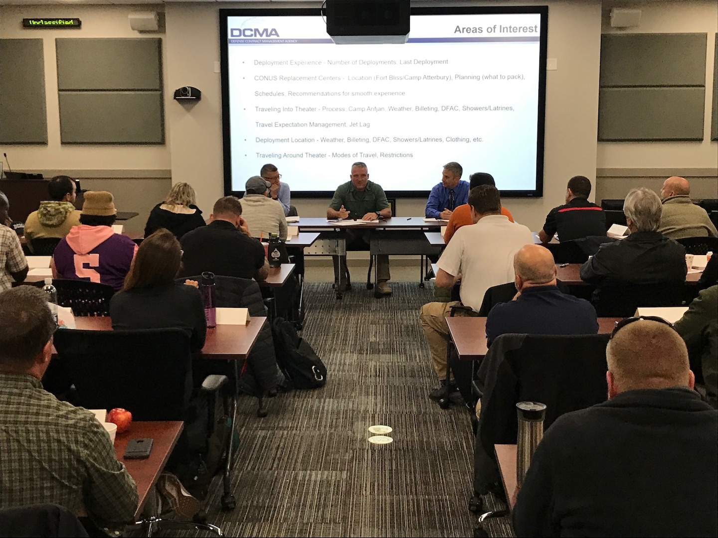 Three men sit at a table talking to other employees in a classroom setting