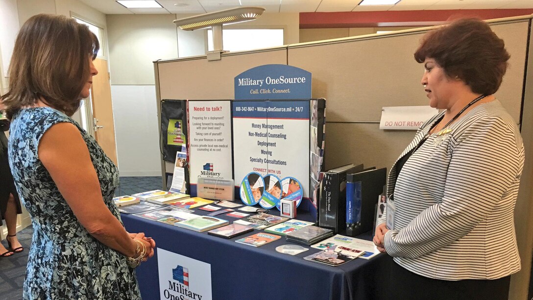 Two women talk in front of a table covered with brochures and signs.