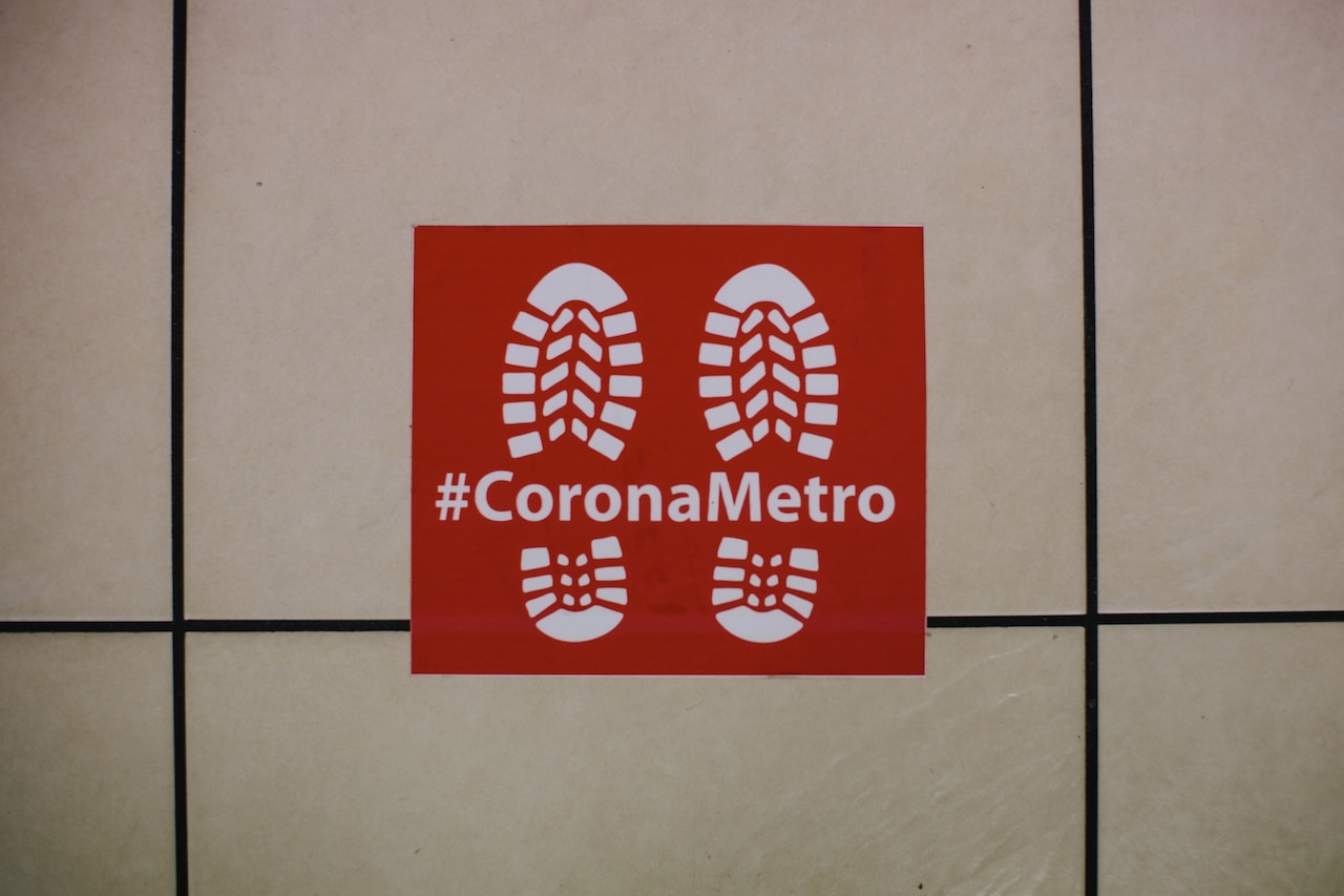 A social distancing sign features the words “Corona Metro” and a pair of combat boot prints on a red background.