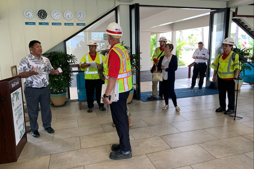 Engineers wearing yellow vests and white hard hats speak with a hotel manager wearing a Hawaiian shirt.