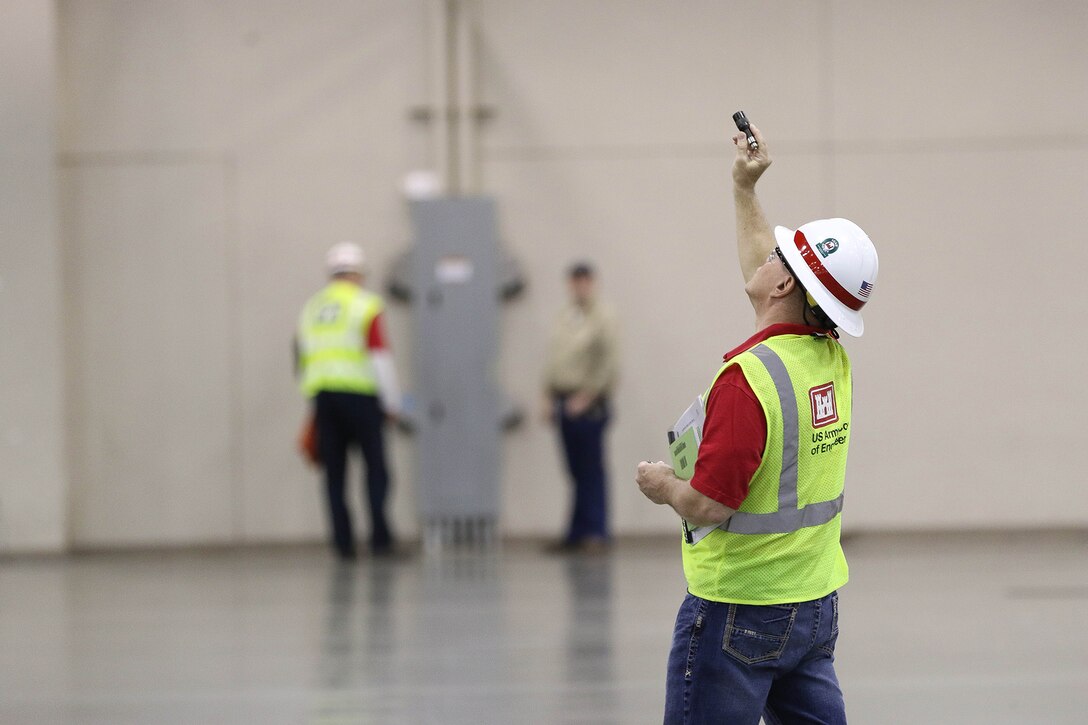 A man wearing a yellow vest and a white hard hat aims a device at a ceiling.