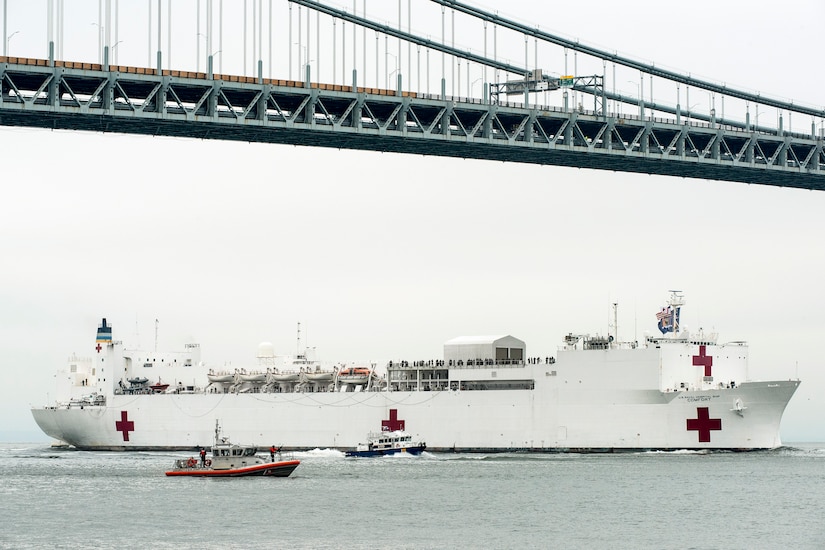 A hospital ship sails under a bridge, accompanied by smaller boats.