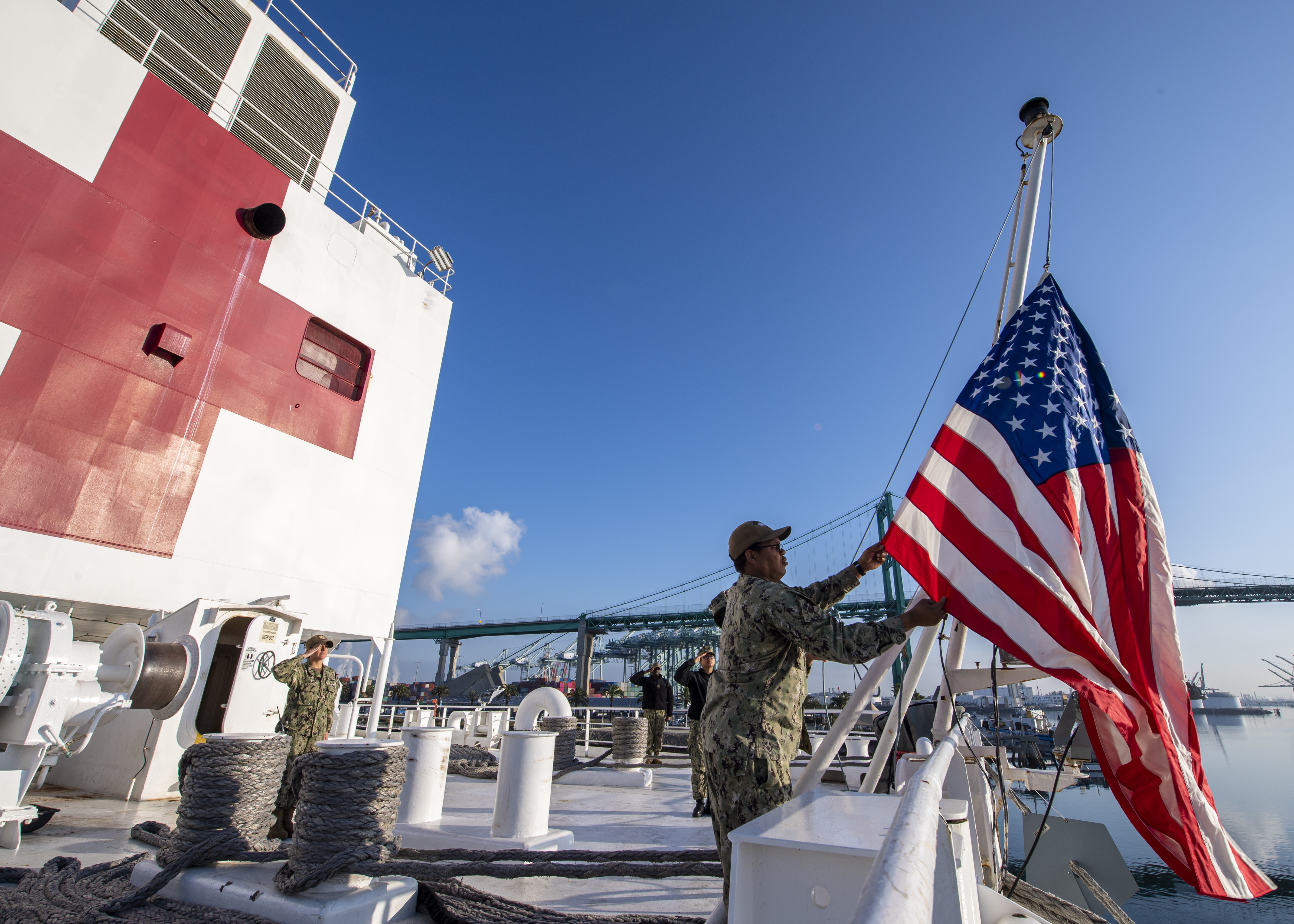 Sailor raises flag on ship marked with a large red cross as another sailor salutes in the background.