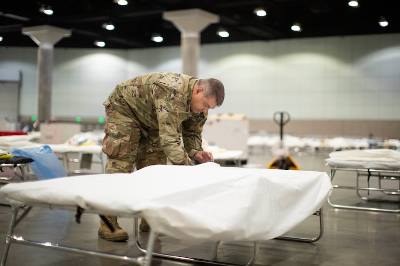 A service member sets up a cot in a large room with many other cots.