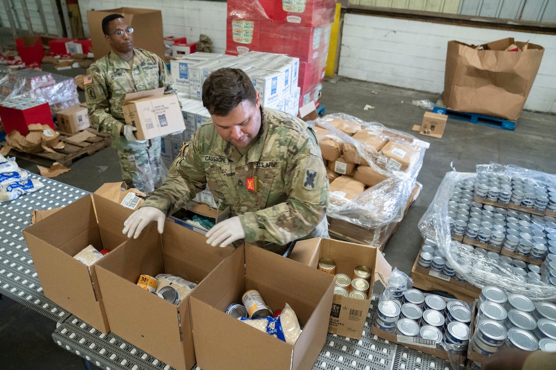 A soldier packs food while another carries a box in the background.