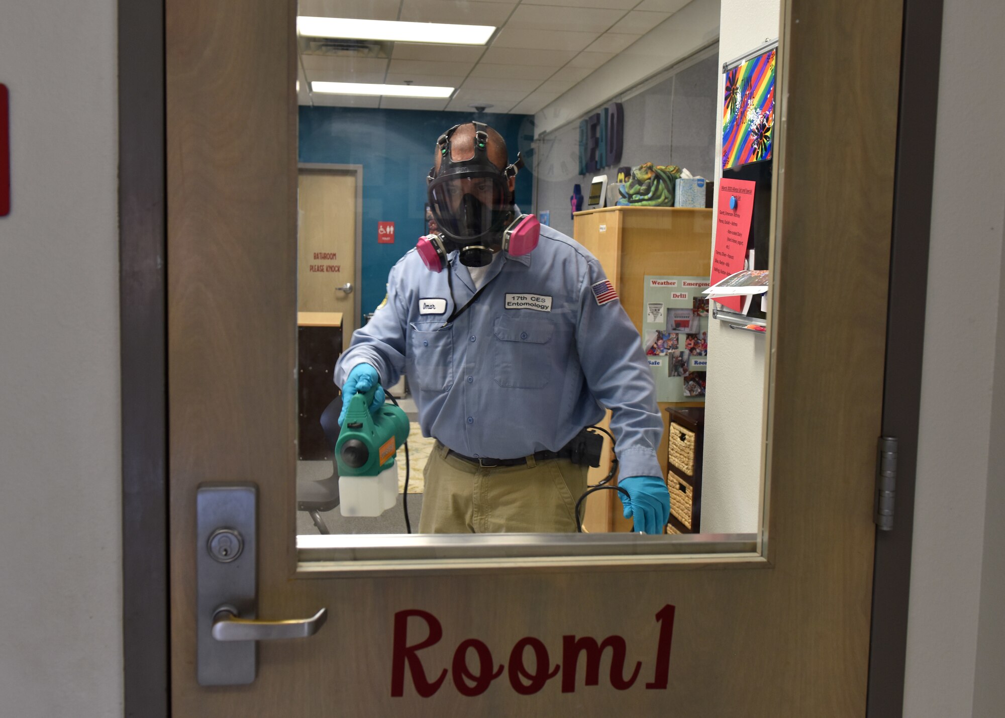 Civil Engineer Squadron Pest Controller Omar Martinez sprays the door to a classroom in the school-age program with disinfectant on Goodfellow Air Force Base, Texas, March 30, 2020. The members of the 17th CES use the fog as a more efficient way to reach every surface in a room. (U.S. Air Force photo by Senior Airman Seraiah Wolf)