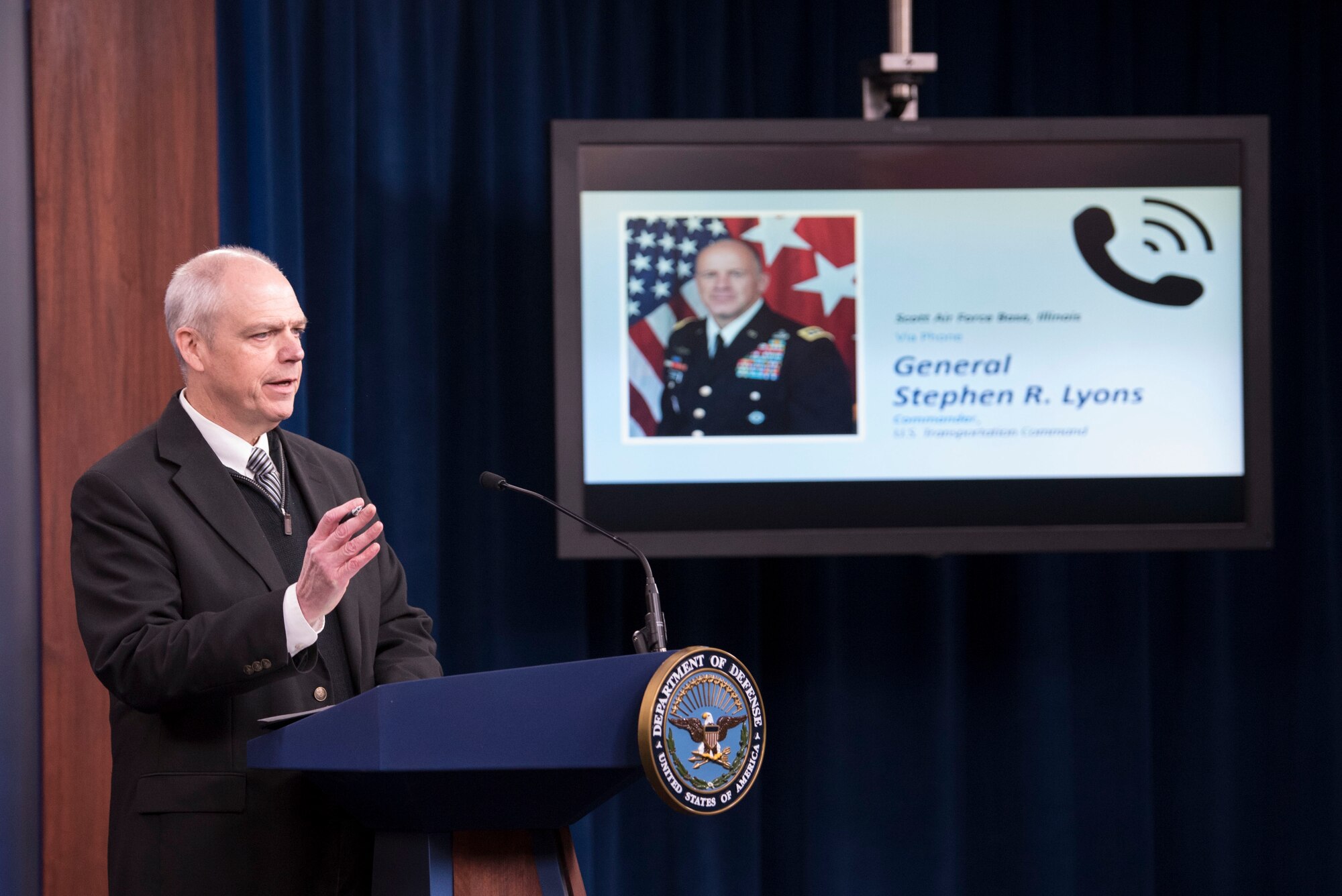 A man stands at a podium with a tv monitor next to him showing a general who is calling in to the room.