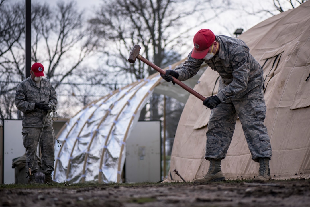 Two service members wearing red hats, face masks and gloves work outside to set up temporary shelters.