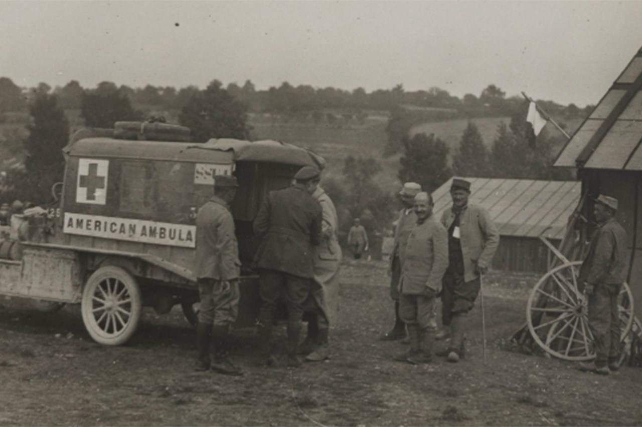 A World War I-era ambulance sits in a field with soldiers milling about.