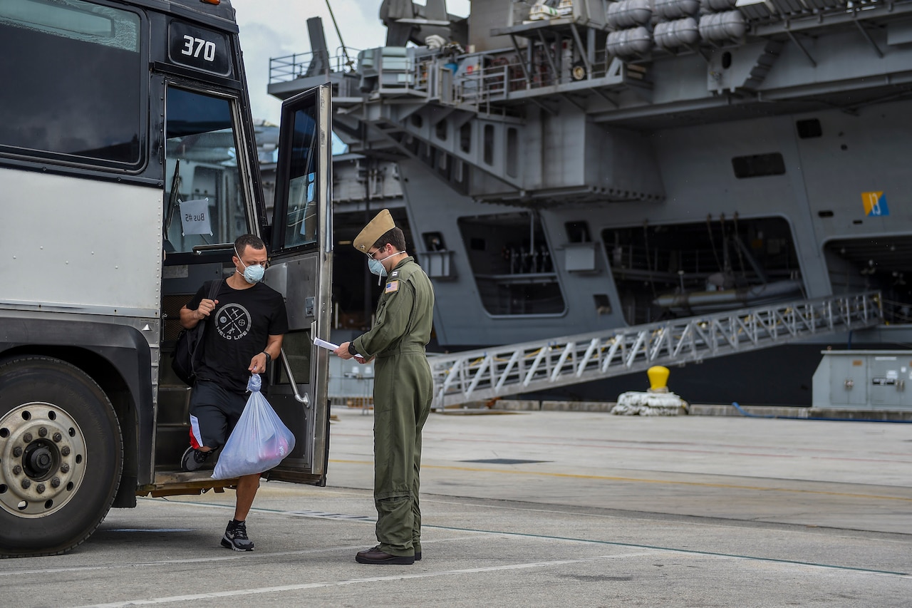 Sailor checks in with officer after getting off the bus. An aircraft carrier is in the background.