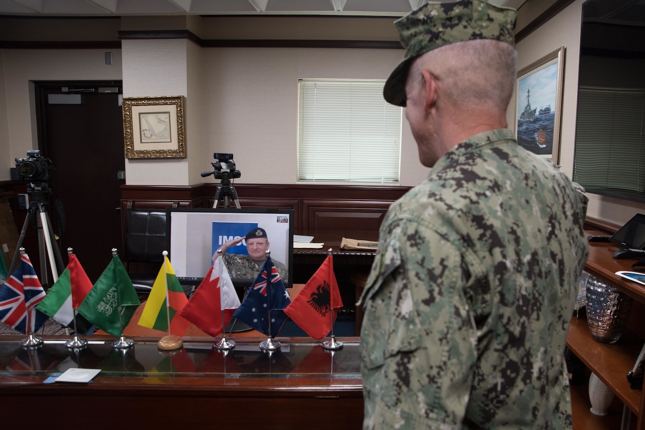 Admiral participates in a virtual change of command as a British naval officer is pictured live on a computer screen. Seven miniature national flags are on the table in front of the computer screen.