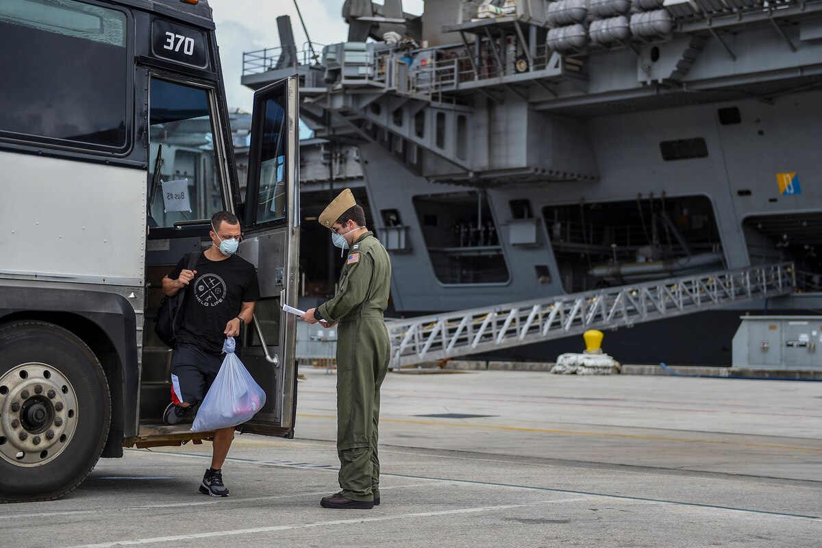 Sailor checks in with officer after getting off the bus. An aircraft carrier is in the background.