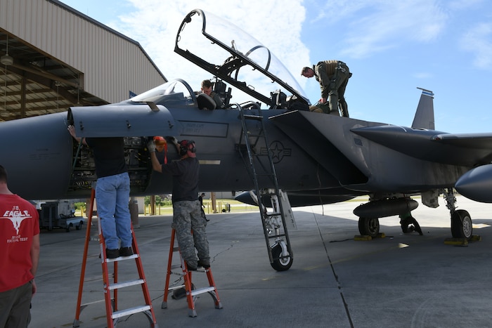 Photo shows two men getting out of an F-15 aircraft using a ladder while two other men on ladders begin working on the nose of the aircraft.