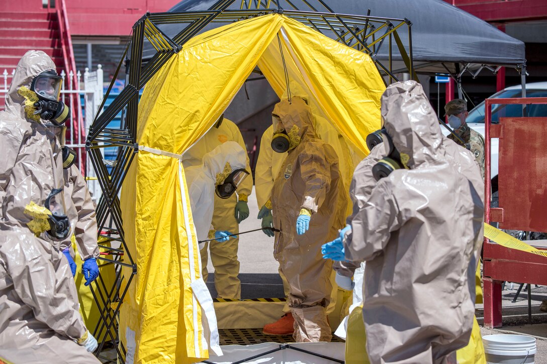 Service members in whole-body protective gear work in and around a yellow tent.