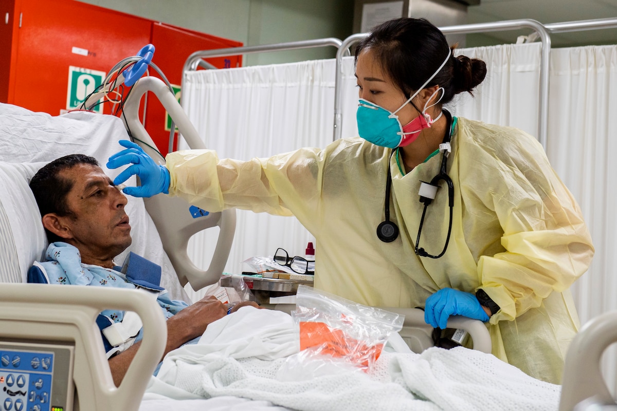 A Navy medic in personal protective equipment removes a bandage from a patient’s face.