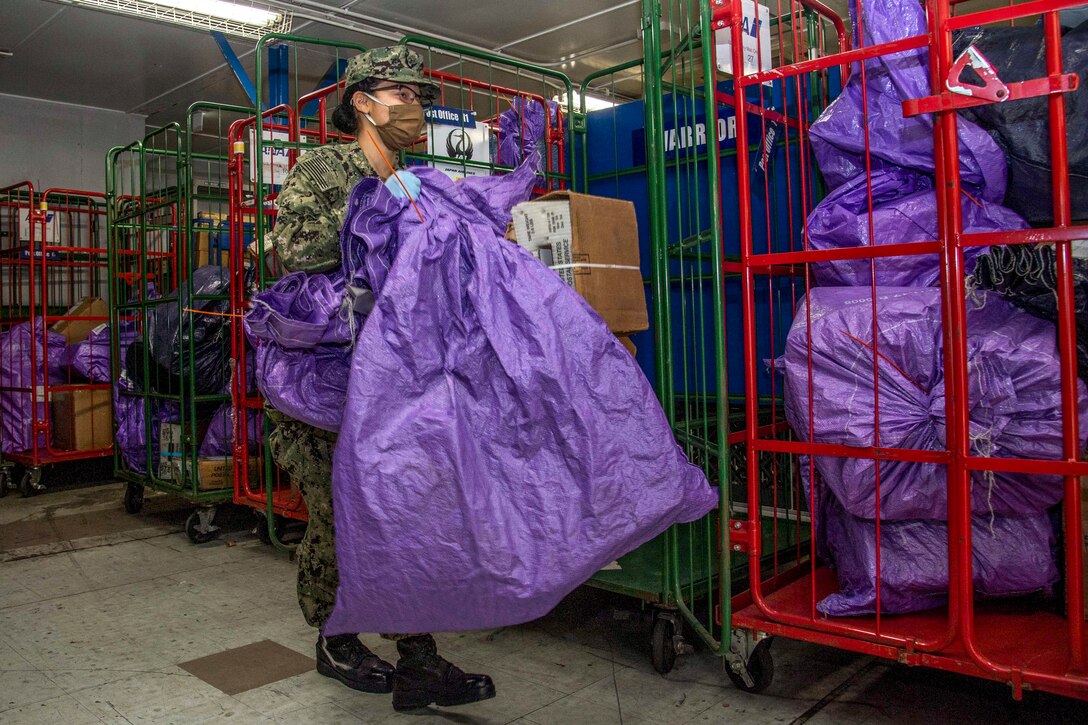 A sailor wearing a mask carries a large purple sack as others sit on wheeled racks nearby.