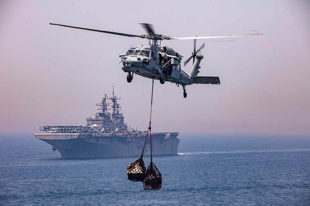 A helicopter carrying supplies flies over open water near a ship.