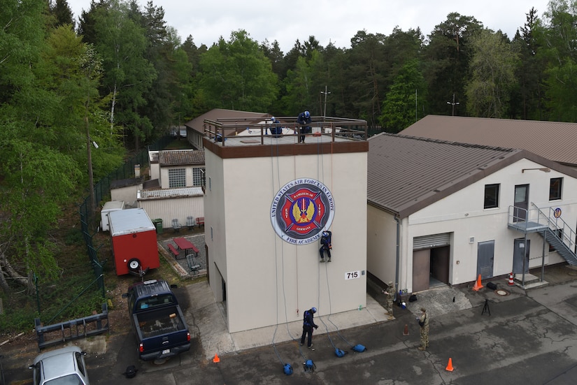 An airman rappels down a building while other airman watch from below and above.