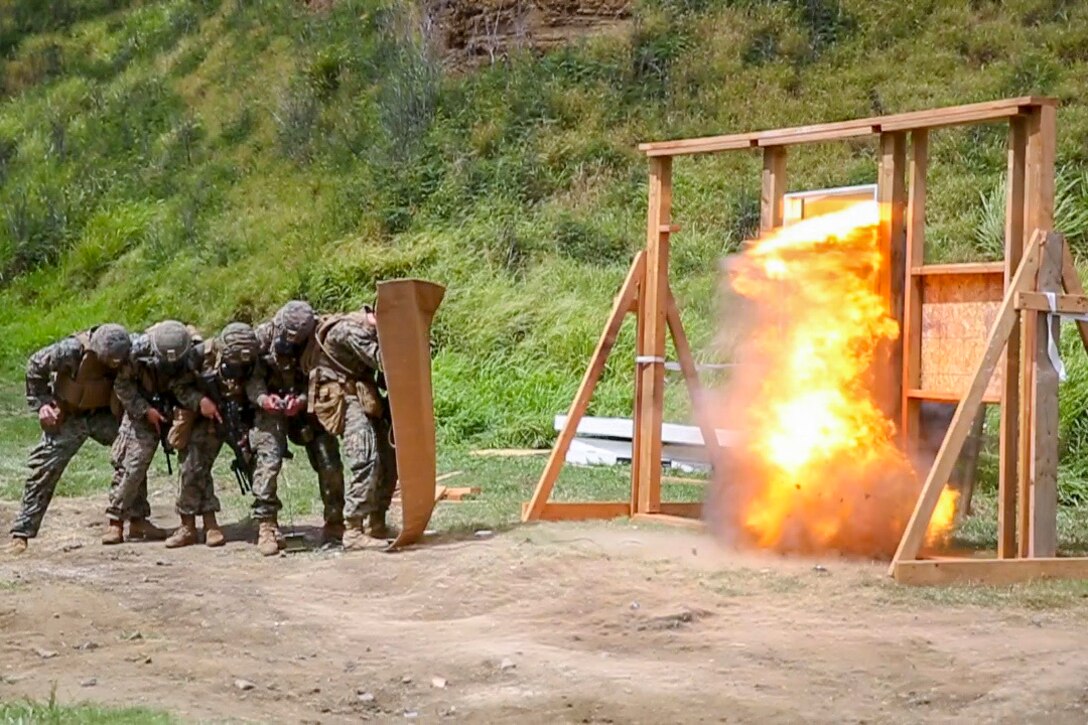 A group of Marines stand behind a barrier as an explosive detonates.