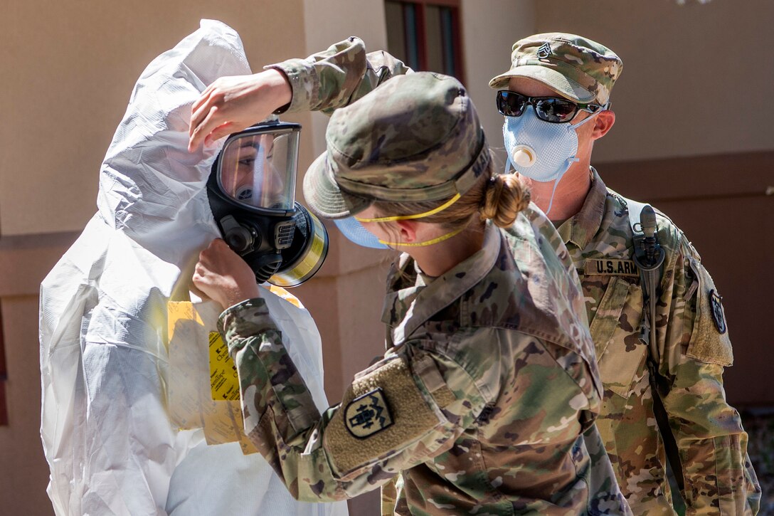 A woman in a military uniform adjusts the face mask of a person wearing a head-to-toe protective suit as another service member observes.