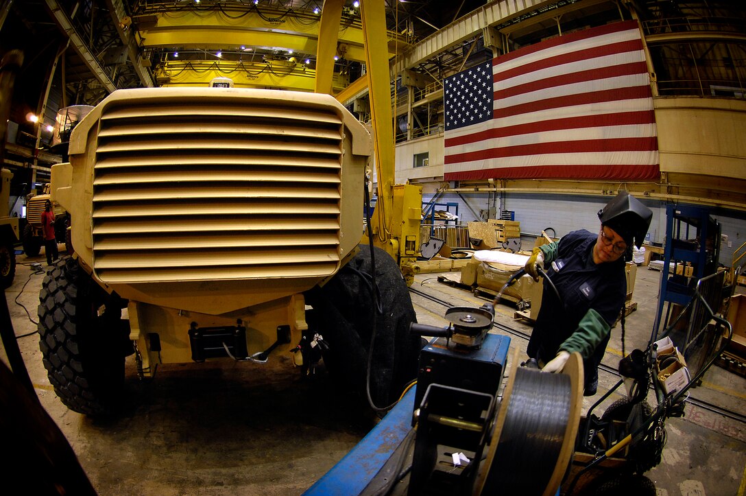 A worker wears a welder’s hood flipped up on her head. She manipulates a roll of wire. A large vehicle sits to her left.