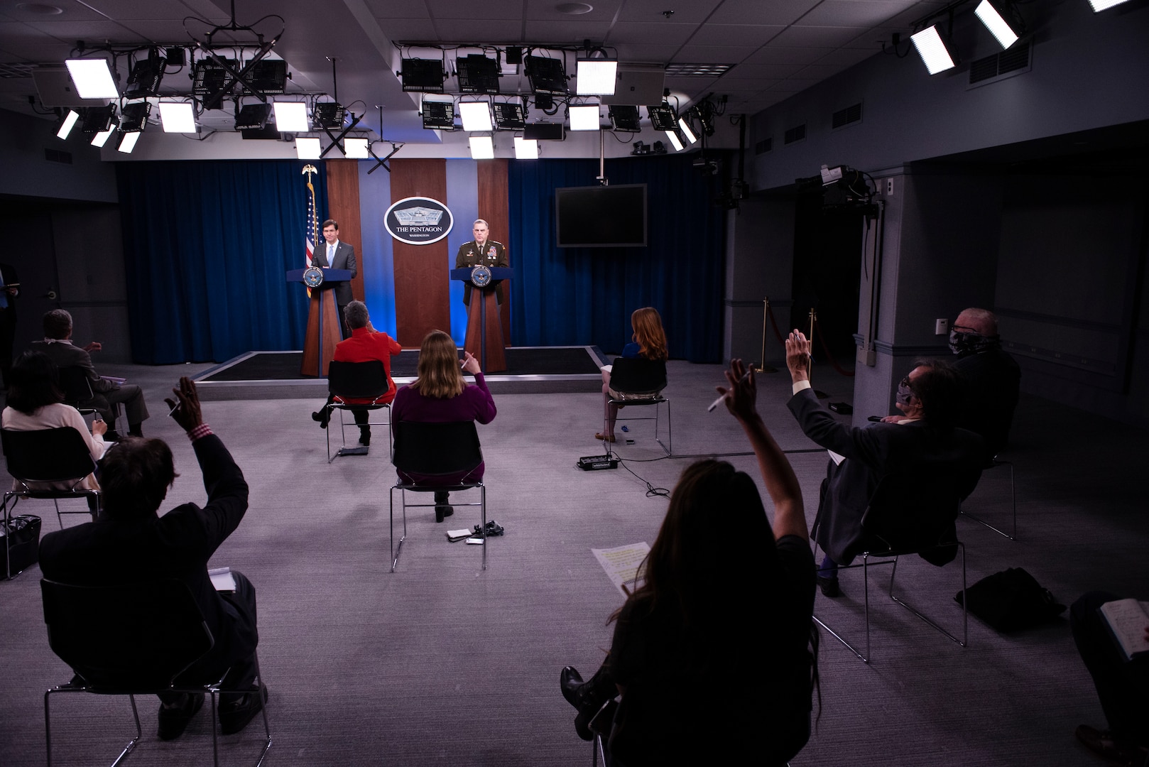 Socially distanced reporters raise their hands at a news conference.